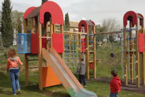 Spielplatz mit rotem Klettergerüst und Rutsche auf dem Campingplatz, Kinder laufen über den Weg im Grünen.