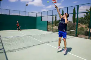 Spieler hebt den Schläger am Tennisplatz, Ball in der Luft, grüner Zaun und Betonwand am Camping-Sportfeld.