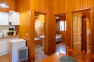 Holzinterieur mit Küchenzeile, Kühlschrank und Heizkörper; dahinter ein Schlafzimmer mit Bett und Türen zum Flur.