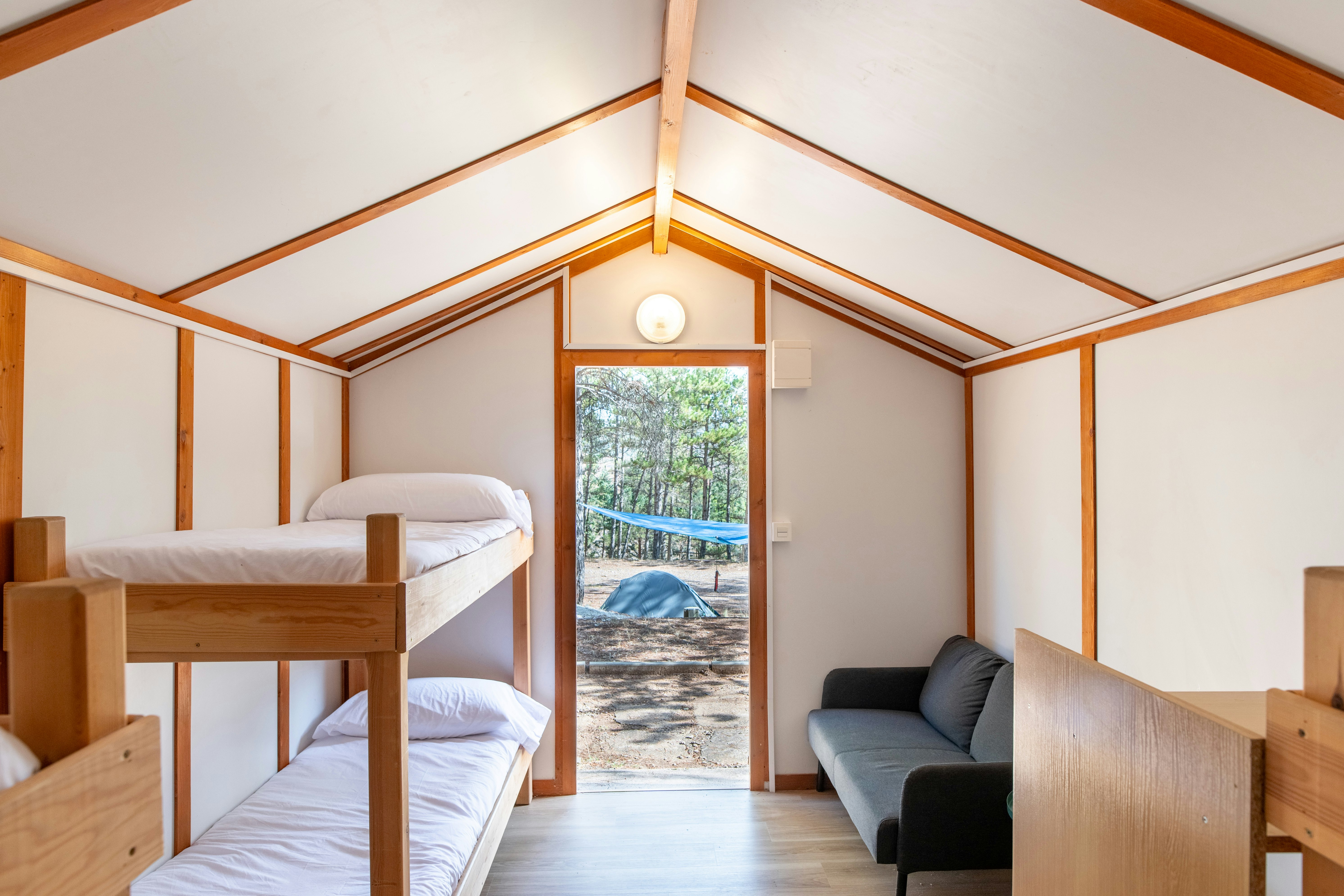 Innenansicht eines Campingzelts mit Etagenbett, Sofa und Holzwänden; Blick durch die Tür auf den Stellplatz und Bäume.