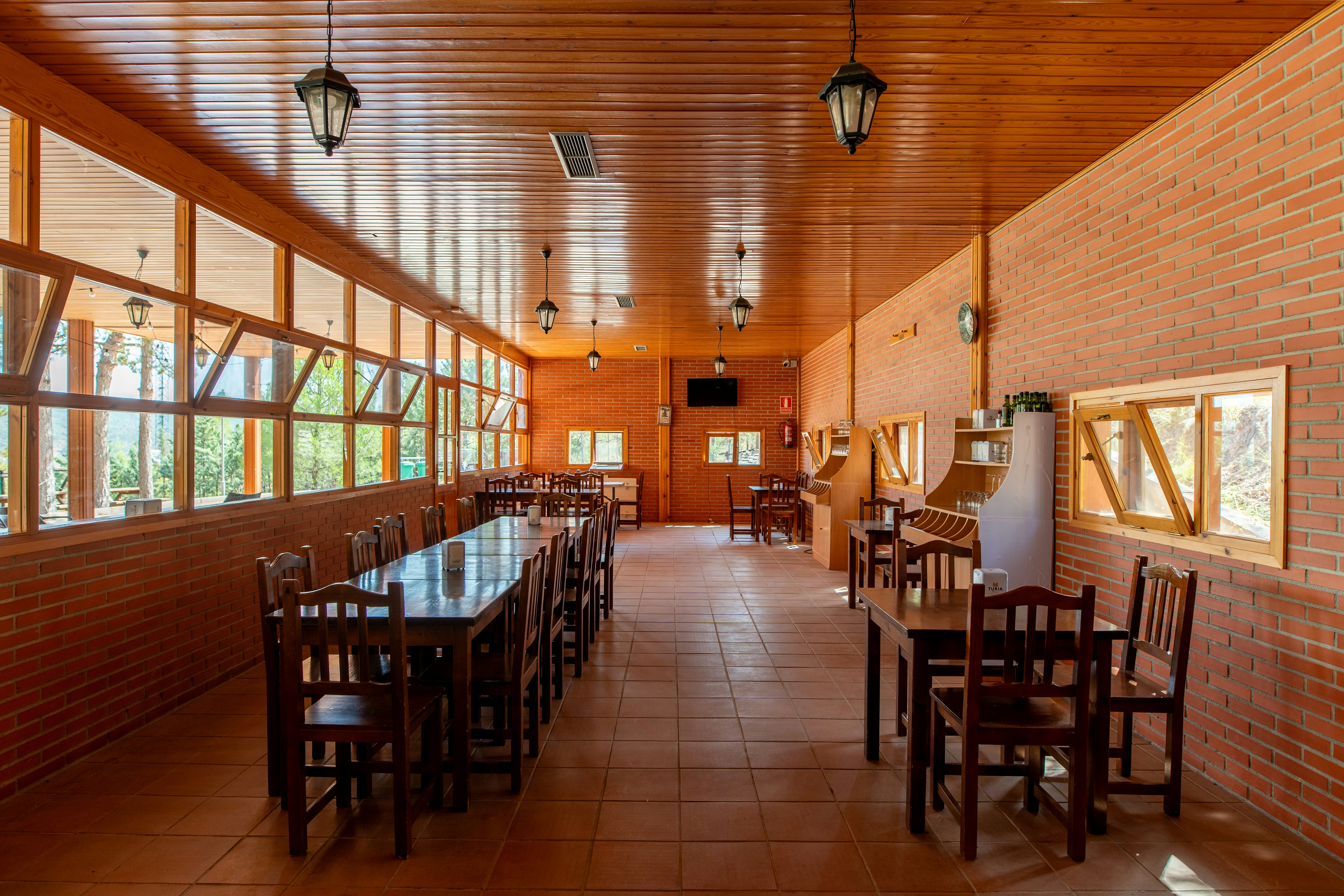 Speisesaal mit langen Holztischen und Stühlen, Ziegelwänden und großen Fenstern, Holzdecke und Lampen im Innenbereich.