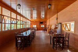Speisesaal mit langen Holztischen und Stühlen, Ziegelwänden und großen Fenstern, Holzdecke und Lampen im Innenbereich.