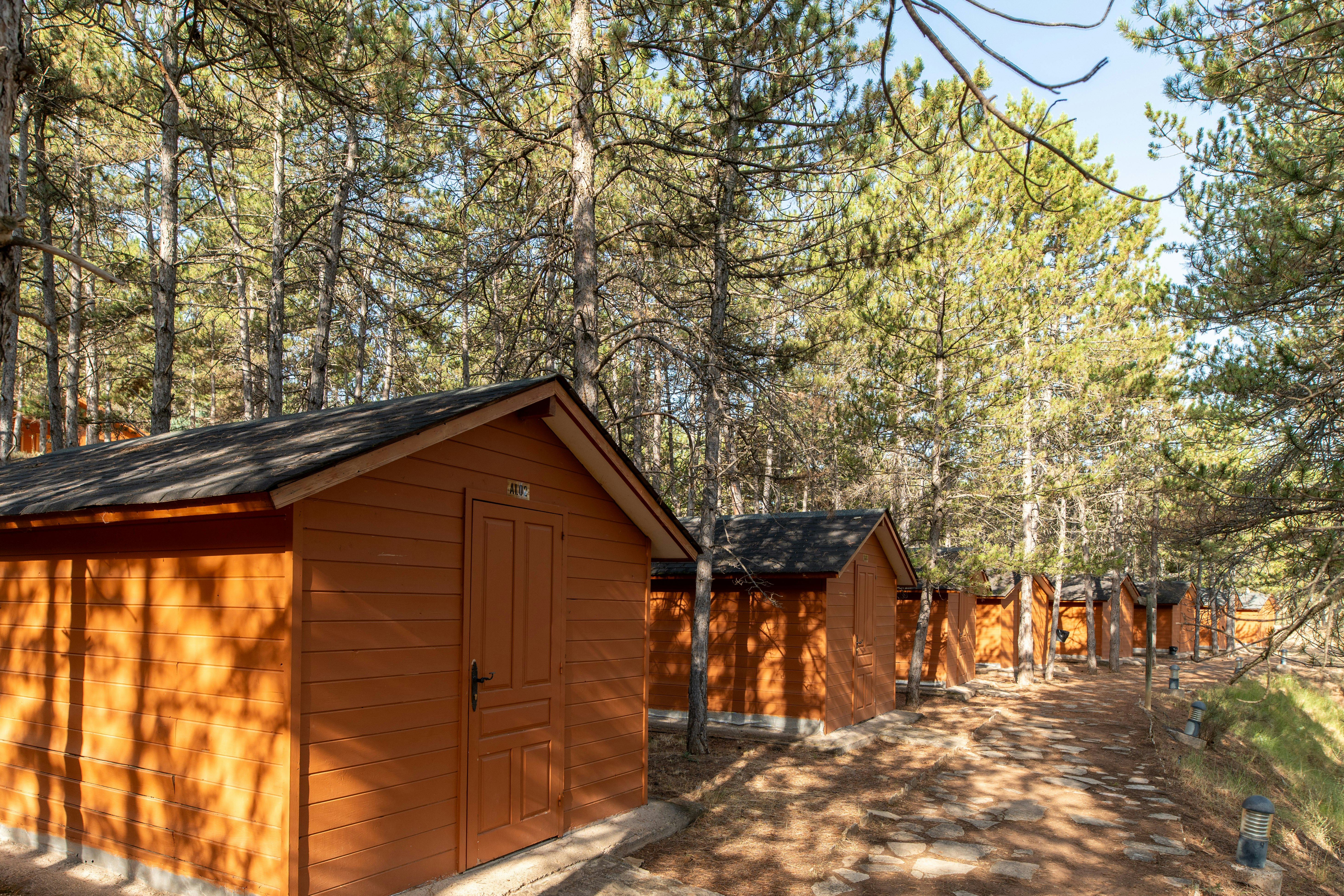 Holzhütte und kleines Nebengebäude auf einem Campingplatz im Wald, mit Holzpfosten-Zaun und Kiesweg.