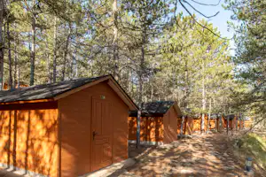 Holzhütte und kleines Nebengebäude auf einem Campingplatz im Wald, mit Holzpfosten-Zaun und Kiesweg.