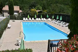 Schwimmbecken mit blauen Fliesen, Liegen und grünen Sonnenschirmen, umgeben von Hecken und Campinggebäuden.