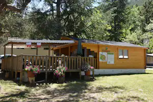 Campingbungalow mit Veranda, Holzgeländer und Blumen vor dem Eingang, umgeben von Bäumen und Rasen.