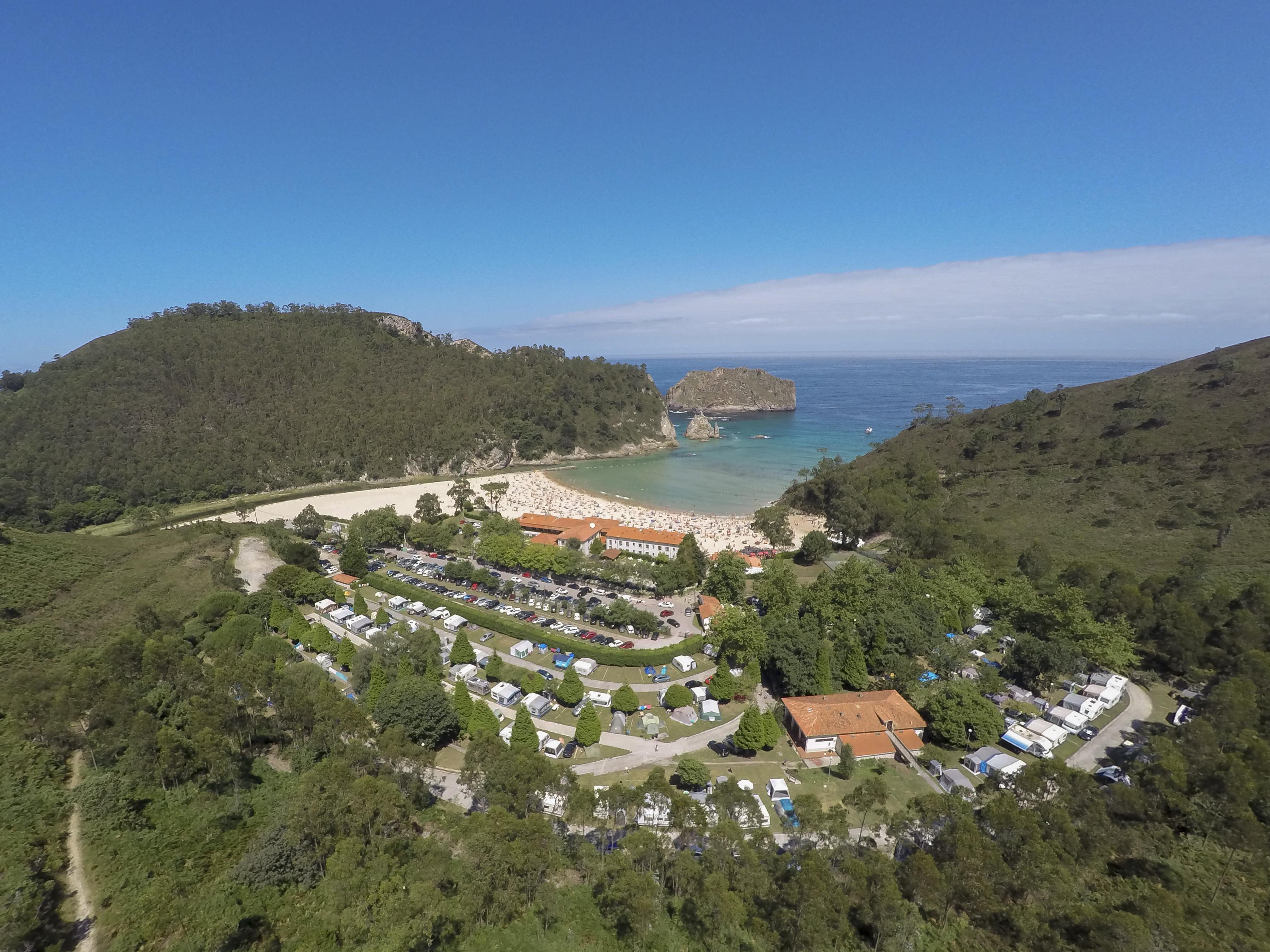 Luftaufnahme eines Campingplatzes mit Wohnwagen und Stellplätzen, umgeben von grünen Hügeln und einer kleinen Bucht am Meer.