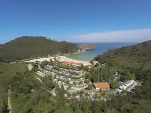Luftaufnahme eines Campingplatzes mit Wohnwagen und Stellplätzen, umgeben von grünen Hügeln und einer kleinen Bucht am Meer.
