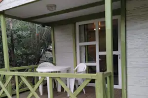 Holzbungalow mit Veranda, Tisch und Stühlen auf der Terrasse, grüne Umrandung und Bäume im Hintergrund.