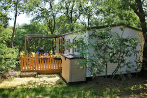 Kleines Mobilheim mit Veranda und Holzgeländer im Grünen, umgeben von Bäumen und Gartenweg.