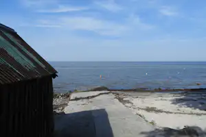 Holzhütte am Campingplatz am Meer, mit Betonweg und Blick auf die Küste und das ruhige Wasser.