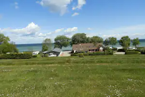 Grüner Campingplatz mit Rasen, Wohnwagen und kleinen Gebäuden am Rand, dahinter das Meer und Bäume.