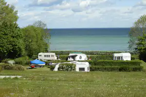 Campingplatz mit Wohnwagen und Zelten auf einer Wiese, Hecken und Bäumen, im Hintergrund das Meer und der Himmel.