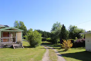 Camping-Bungalow mit Holzterrasse, Rasenfläche und Kiesweg, umgeben von Bäumen und Sträuchern im Garten.