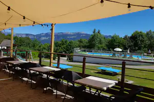 Terrasse mit Tisch und Stühlen unter Sonnensegel, Blick auf den Pool, Liegen und Berge hinter Bäumen.