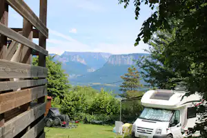 Wohnmobil auf dem Campingplatz mit Blick auf Berge, umgeben von Bäumen und einer Holzterrasse.