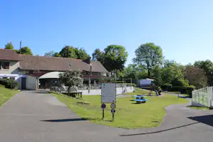 Campinggebäude mit Rezeption und Bar-Schild, Terrasse mit Stühlen, Spielbereich und Rasenfläche zwischen Bäumen.