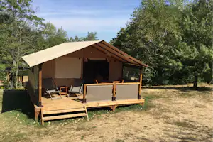 Campingzelt auf Holzplattform mit Markise, zwei Liegestühlen und Sichtschutz, umgeben von Bäumen im grünen Campingbereich.