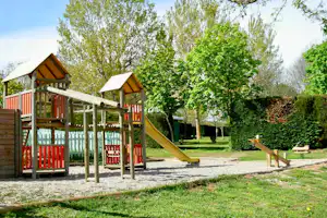 Spielplatz mit Klettergerüst und Rutsche, rotem Holzgeländer, Kiesweg und Bäumen im grünen Campingbereich.