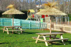 Grüner Campinggarten mit Picknicktischen und Strohdächern, Palmen und Spielgeräten hinter einem weißen Zaun.