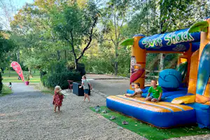 Bunte Hüpfburg „Surf Shack“ auf dem Campingplatz, Kinder spielen auf dem blauen Sprungkissen neben Kiesweg und Bäumen.