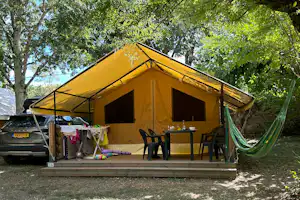 Gelbes Cabana-Zelt auf Holzterrasse mit Tisch und Stühlen, davor ein grüner Hängematten-Stoff und Fahrzeuge im Hintergrund.