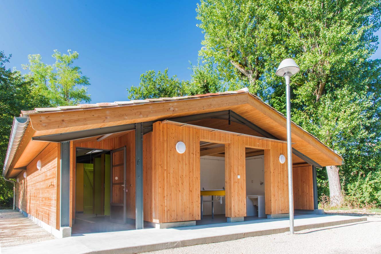 Holz-Blockhaus mit überdachter Terrasse, Eingangstür und großen Fenstern, daneben Laterne und Weg vor Bäumen.