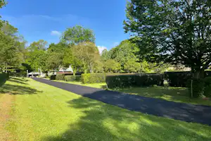 Asphaltweg im Campinggelände, umgeben von Rasen, Hecken und Bäumen, mit kleinen Gebäuden im Hintergrund.