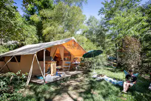 Campingzelt mit Vorzelt im grünen Garten, Tisch und Stühle daneben, daneben sitzt eine Person auf einer Decke.