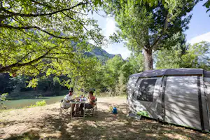 Campingzelt am Flussufer im Grünen, daneben Tisch und Stühle unter Bäumen, im Hintergrund Berge und Wasser.