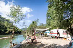 Campingplatz am Fluss mit Wohnwagen, Wäscheleine und Sitzplatz am Ufer, Bäume und Berge im Hintergrund.