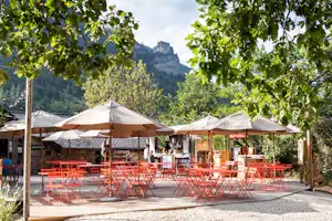 Terrasse eines Campingplatzes mit roten Tischen und Stühlen unter Sonnenschirmen, umgeben von Bäumen und Bergen im Hintergrund.