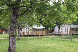 Campingbungalows mit Holzterrassen im Grünen, umgeben von großen Bäumen und Rasenfläche.