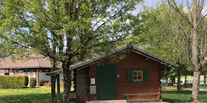 Holzhütte mit grüner Tür und Fenster, davor Bank und Weg, umgeben von Bäumen und Rasen auf dem Campingplatz.
