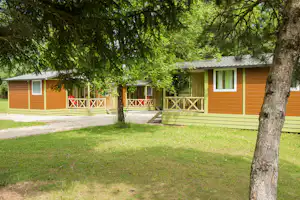 Bungalow mit Veranda und Holztreppe, umgeben von Bäumen und grünem Rasen auf dem Campingplatz.