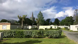 Reihe von Wohnmobilen und Mobilheimen auf einem Campingplatz, Hecken und Rasen sowie ein Weg zur Kirche im Hintergrund.