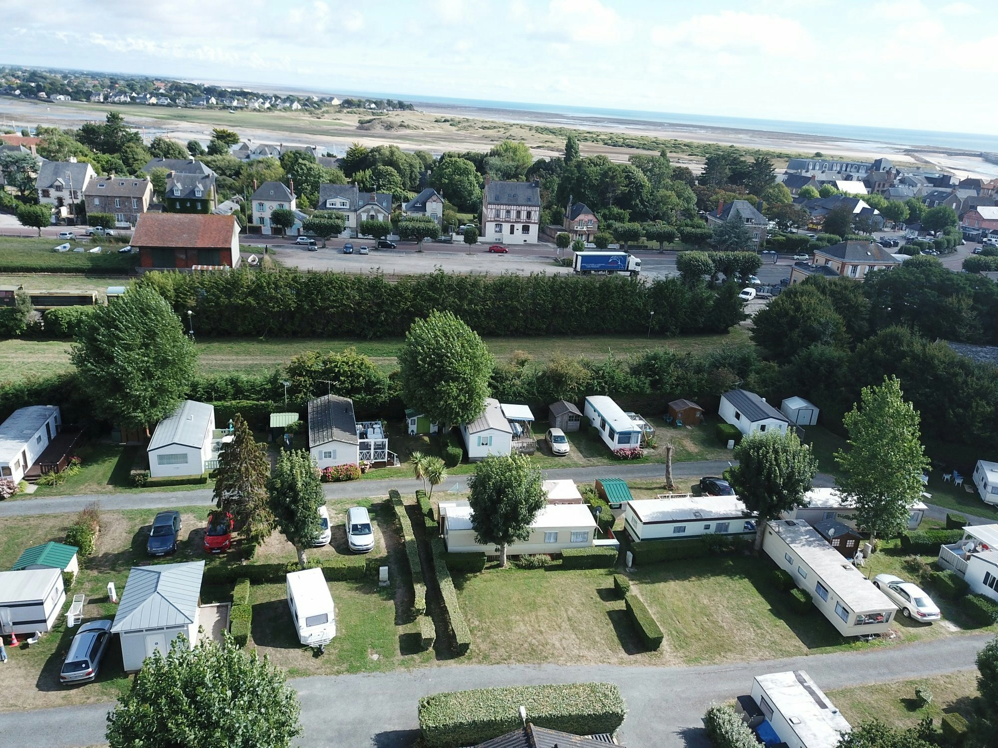 Campingplatz mit Wohnwagen und Zelten auf Stellplätzen, umgeben von Bäumen, Blick auf Häuser und die Küste in der Ferne.