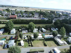 Campingplatz mit Wohnwagen und Zelten auf Stellplätzen, umgeben von Bäumen, Blick auf Häuser und die Küste in der Ferne.