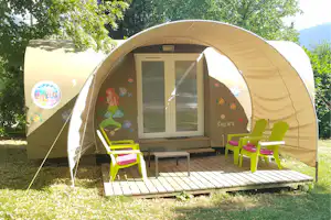 Familienzelt mit Kinderdekor, Glastür und zwei Fenstern; Holzterrasse mit bunten Stühlen im Grünen.