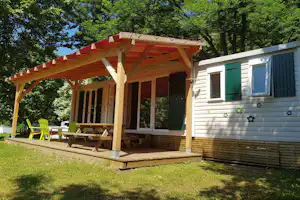 Mobilheim mit Holzveranda, großen Fenstern und grünen Türen, davor Holzterrasse mit Stühlen im Grünen.