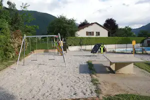 Spielplatz auf dem Campingplatz mit Sandfläche, Schaukeln, Klettergerüst und Terrasse neben einem weißen Haus im Grünen.