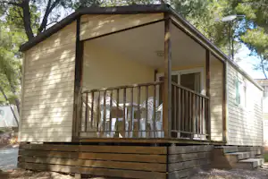 Wohnwagen mit Veranda und Holzgeländer im Grünen, seitliche Fenster und Eingangstür am Campingplatzgebäude.