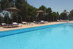 Blauer Campingpool mit mehreren weißen Liegen und Sonnenschirmen auf der Terrasse, umgeben von Bäumen und Sichtschutzzaun.