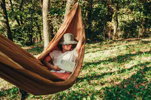 Person in einer Hängematte im Wald, liest ein Buch, umgeben von Bäumen und grünem Unterholz.