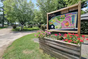 Holztafel mit Campingplatz-Plan, Blumenbeet im Vordergrund, Kiesweg und Rasenfläche zwischen Bäumen auf dem Gelände.