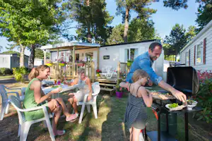 Familie beim Grillen am Campingplatz vor einem Wohnwagen, mit Gartenmöbeln, Kindern und Bäumen im Hintergrund.
