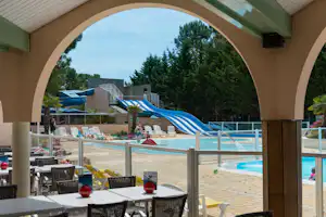 Blick durch einen Torbogen auf den Poolbereich mit Liegen, Sonnenschirmen und Tischen, im Hintergrund Bäume und Gebäude.