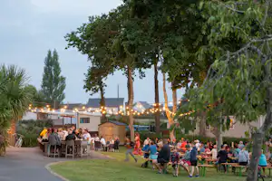 Gartenbereich mit langen Tischen und vielen Campern, beleuchtete Bäume und Wege am Campingplatz, im Hintergrund Gebäude.