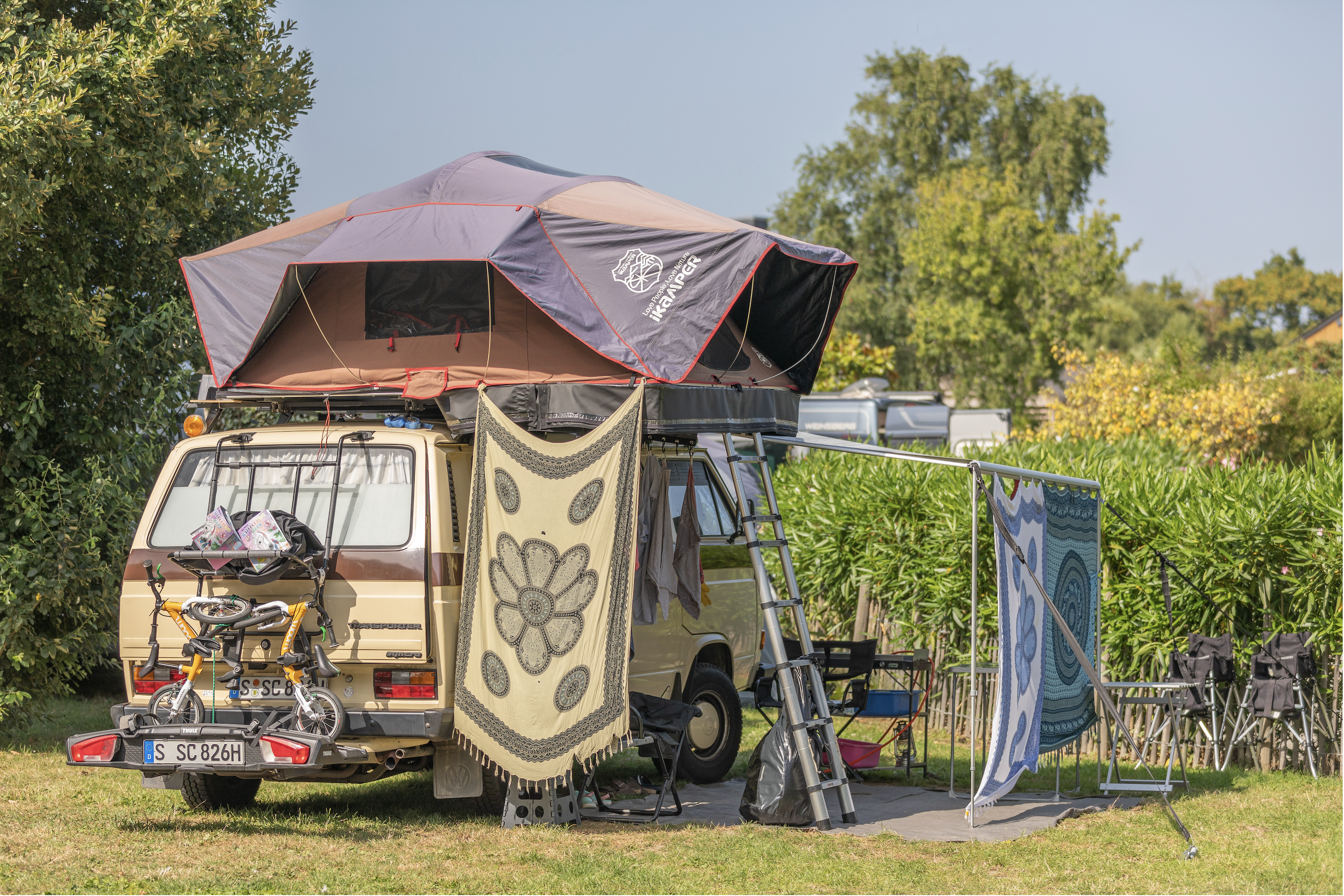 Wohnmobil mit Vorzelt und Markise auf einem Campingplatz, davor Stühle und Tisch, im Hintergrund Hecken und Bäume.