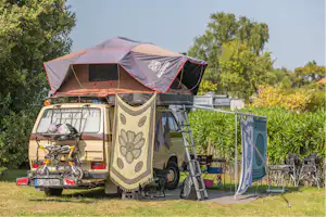 Wohnmobil mit Vorzelt und Markise auf einem Campingplatz, davor Stühle und Tisch, im Hintergrund Hecken und Bäume.